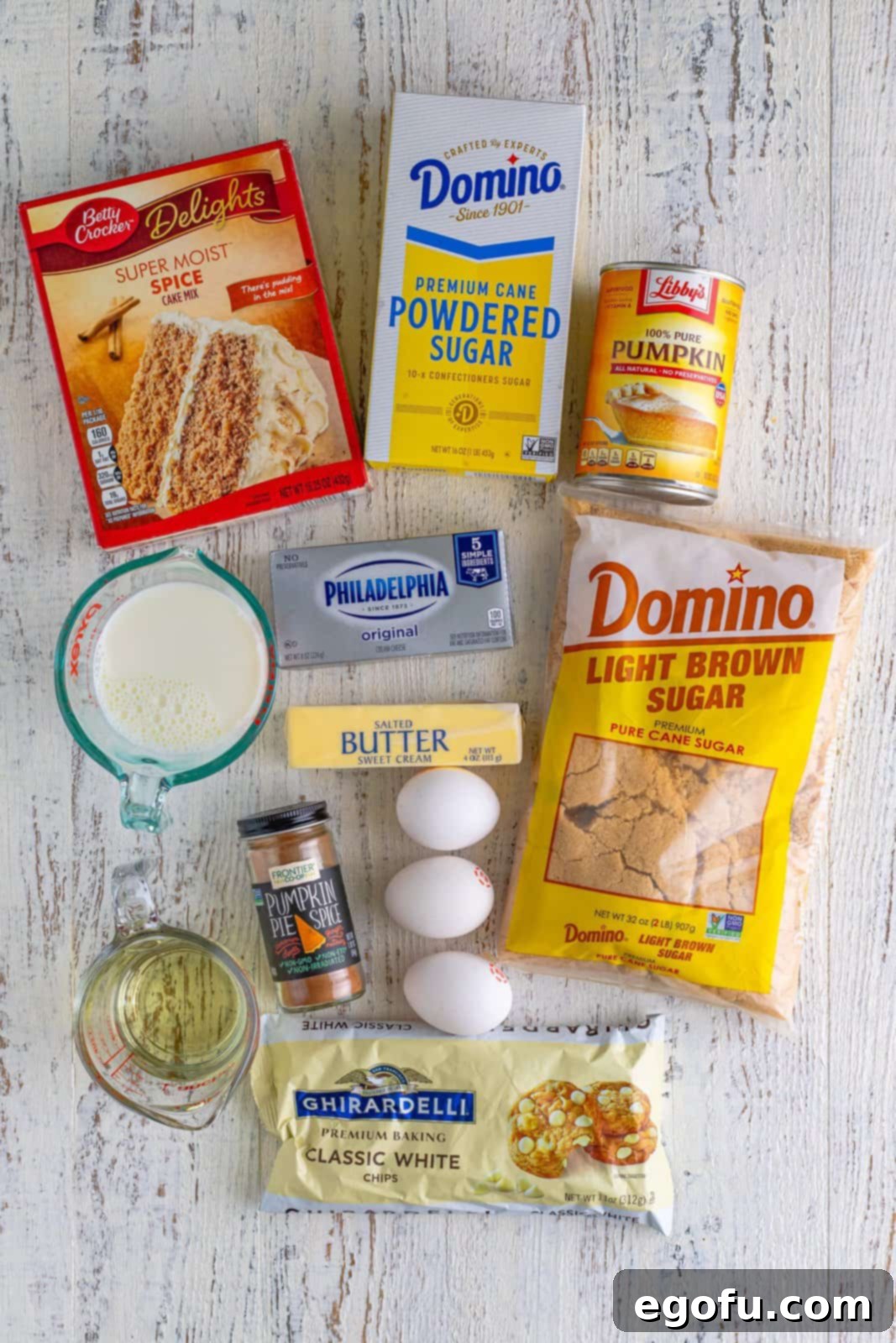 All ingredients laid out on a table: spice cake mix, pure pumpkin puree, milk, eggs, vegetable oil, light brown sugar, cream cheese, salted butter, powdered sugar, pumpkin pie spice and white chocolate chips.