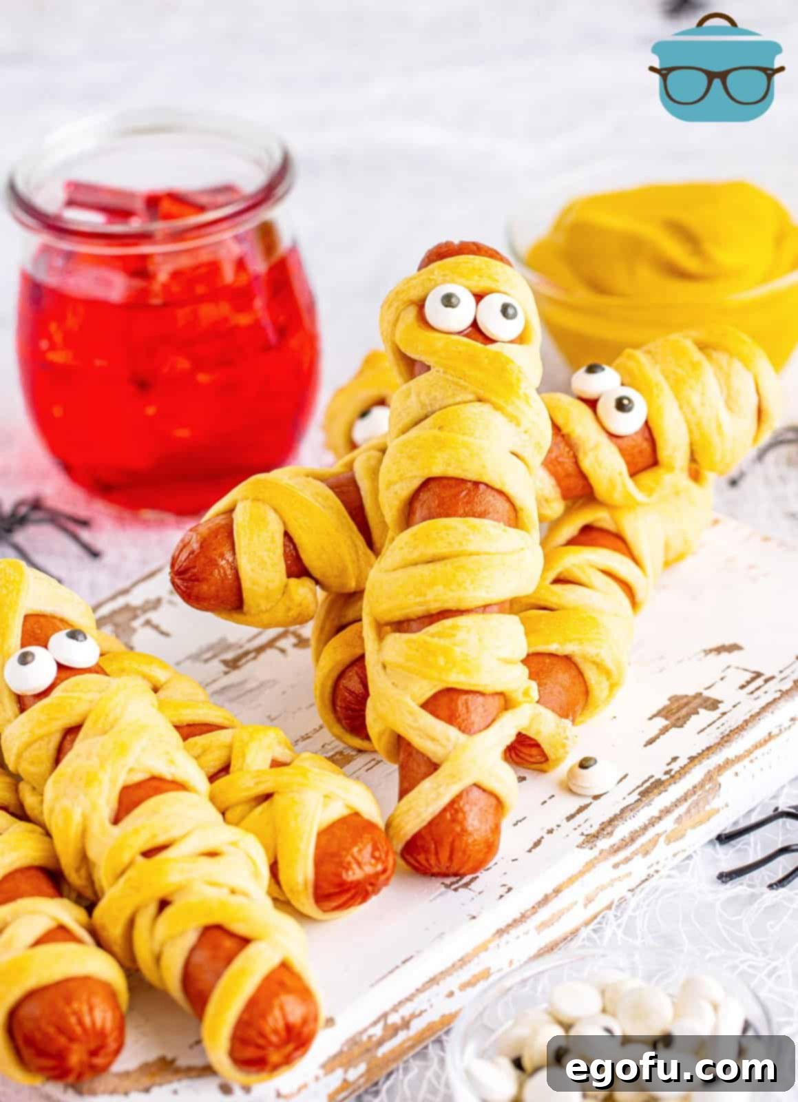 Festive Halloween Mummy Dogs arranged on a white wooden board with a bottle of ketchup in the blurred background, perfect for a spooky snack or meal.