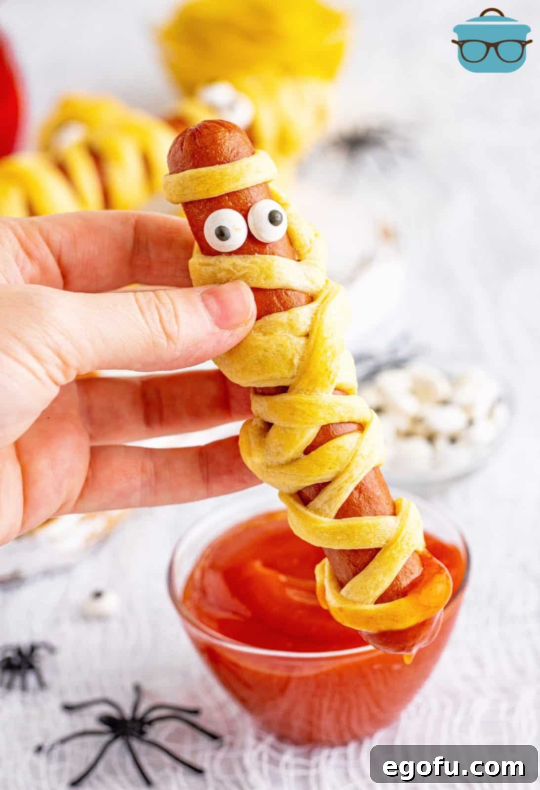 A hand holding up one perfectly baked Halloween Mummy Dog, showcasing its golden crust and candy eyes.