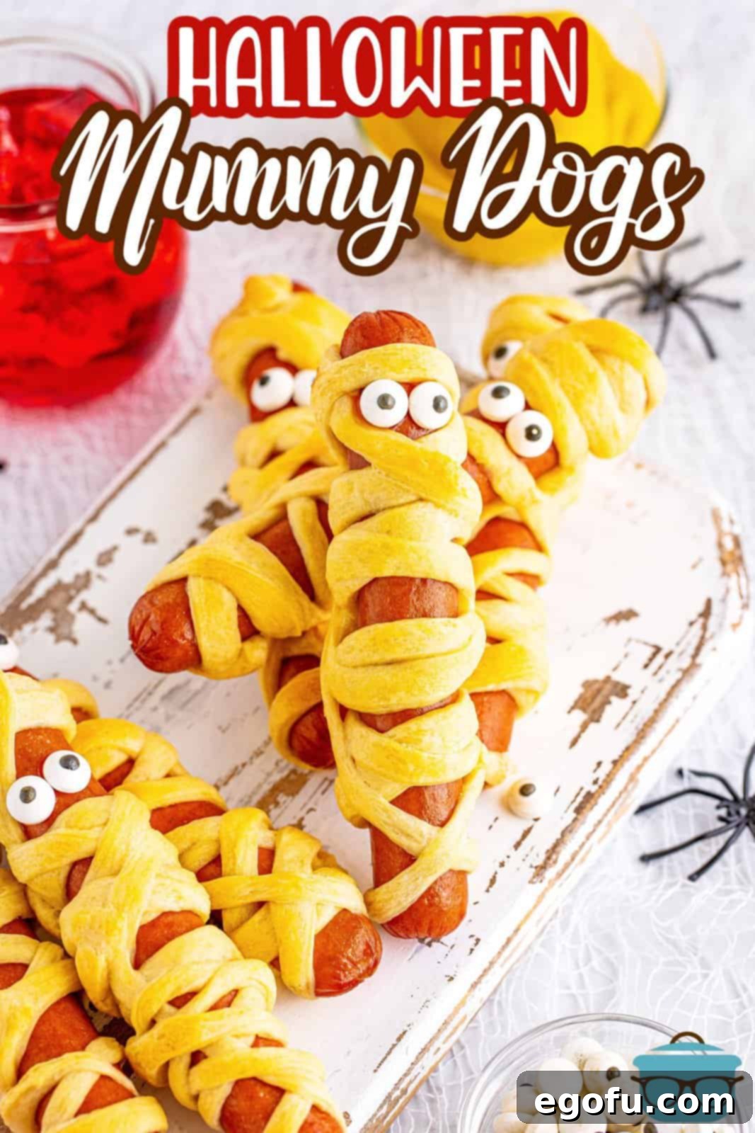 Overhead view of perfectly baked and stacked Halloween Mummy Dogs on a white wooden board with a bottle of ketchup visible in the background.
