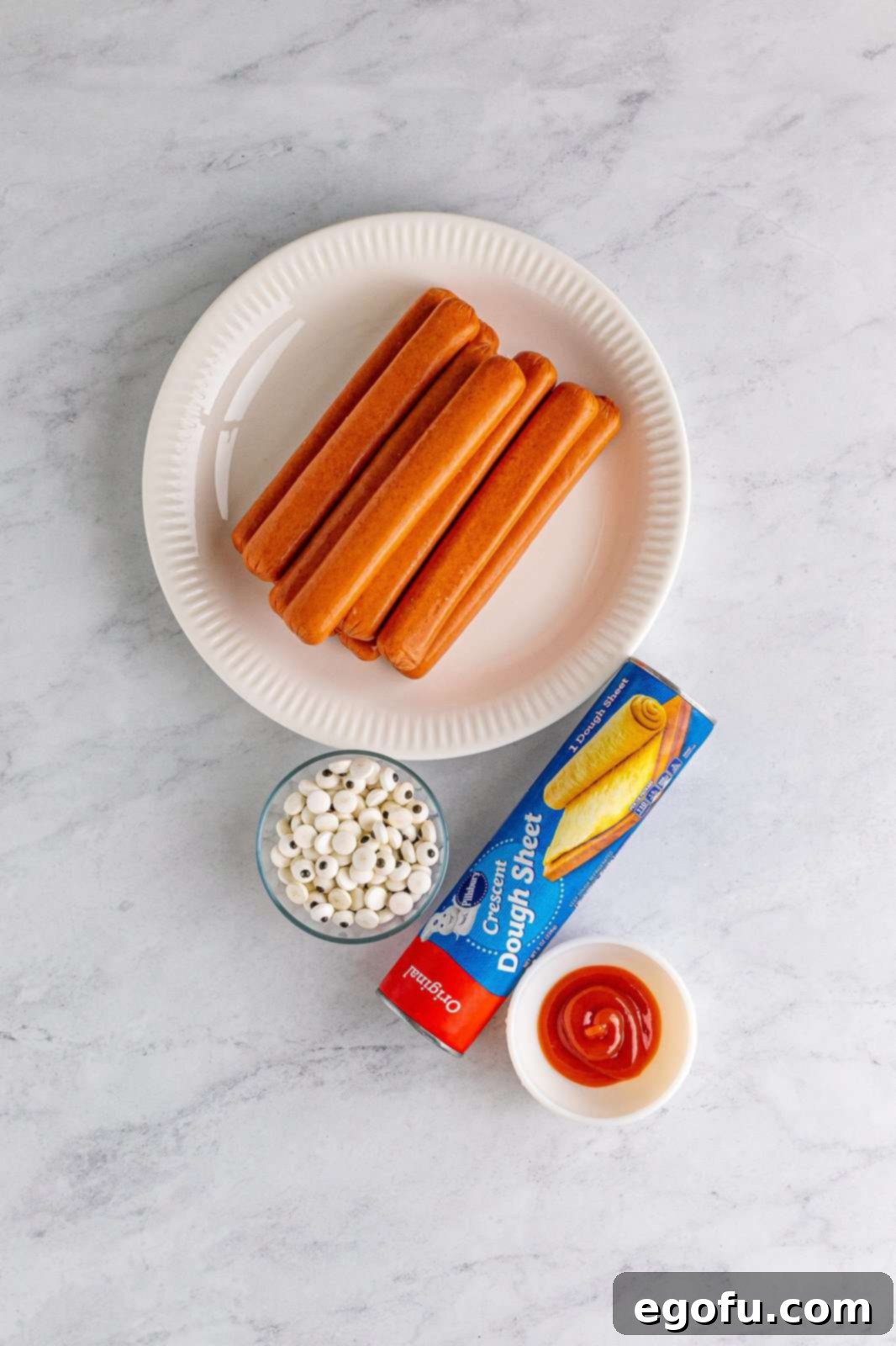 All ingredients laid out: hot dogs, a sheet of crescent roll dough, ketchup, and candy eyes, with various dipping sauces in the background.
