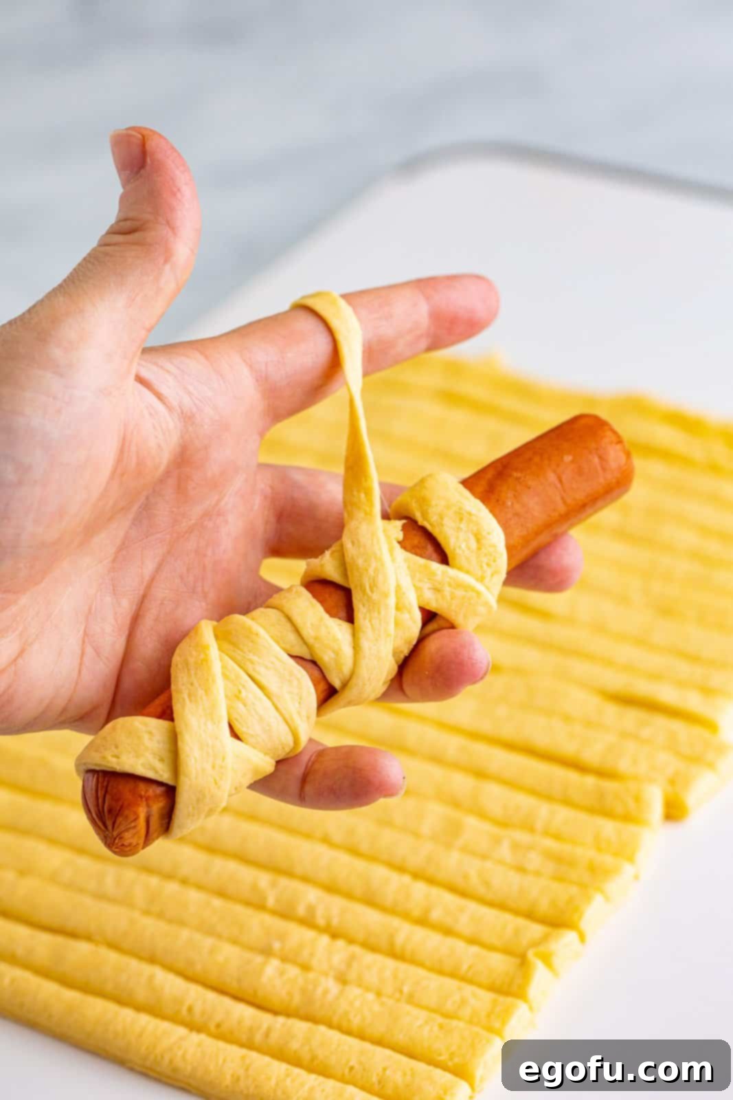 A hand expertly wrapping a hot dog with crescent dough strips, creating the mummy effect.