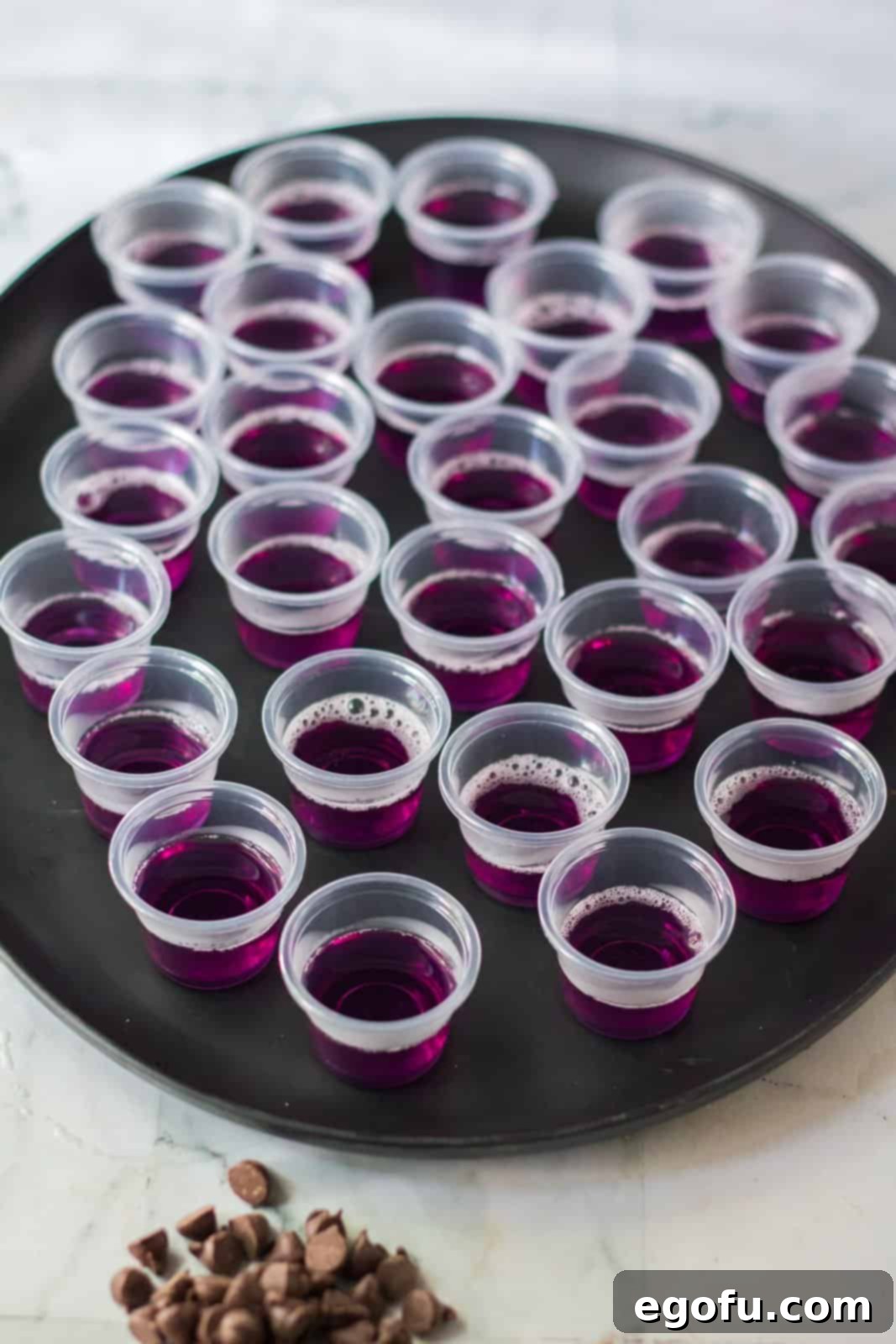 The vibrant purple Jell-O mixture meticulously poured into small plastic glasses, creating a uniform first layer and neatly arranged on a tray, ready for chilling.