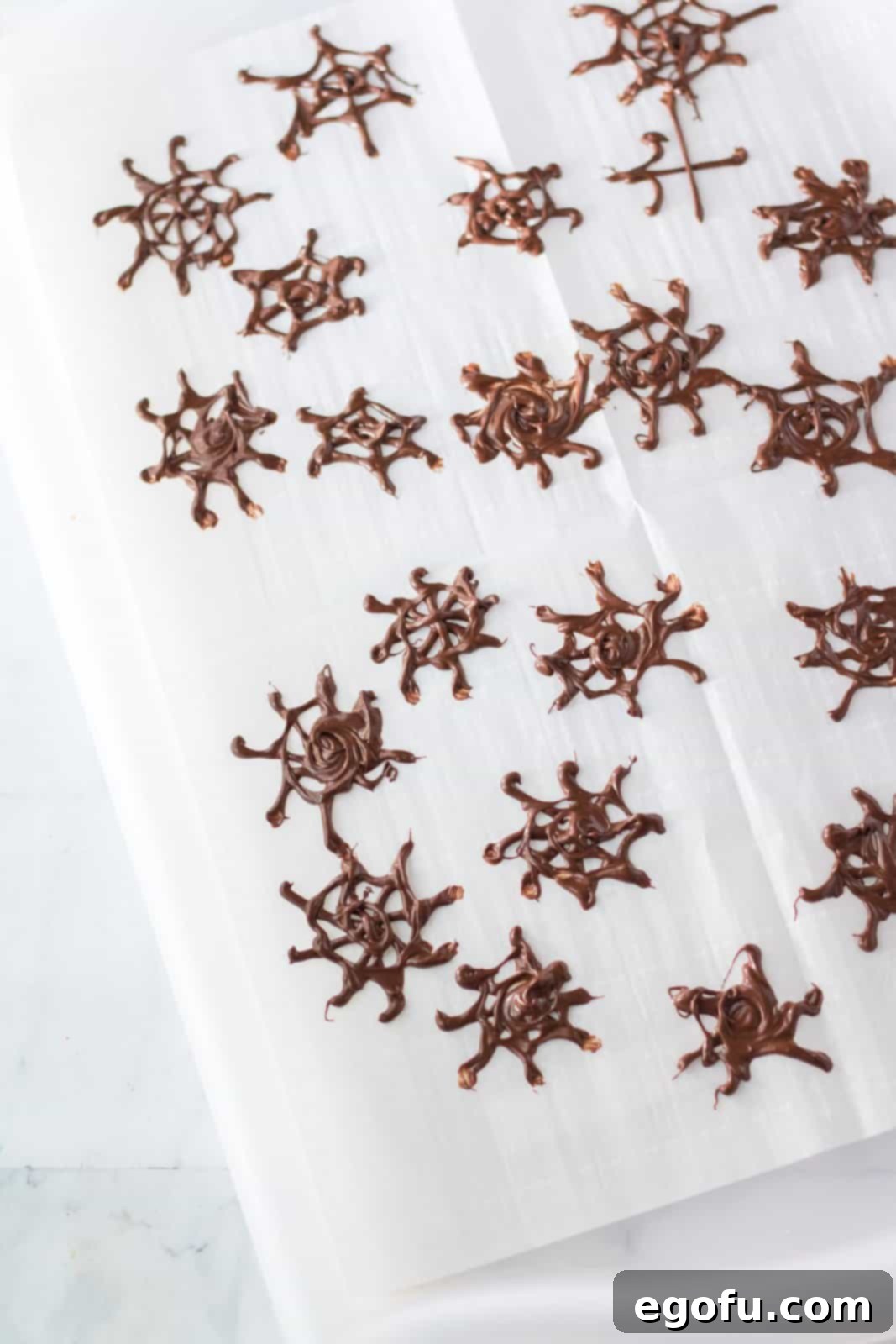 Intricate chocolate spiderwebs delicately piped onto parchment paper, ready to be chilled and hardened for a spooky Halloween decoration.