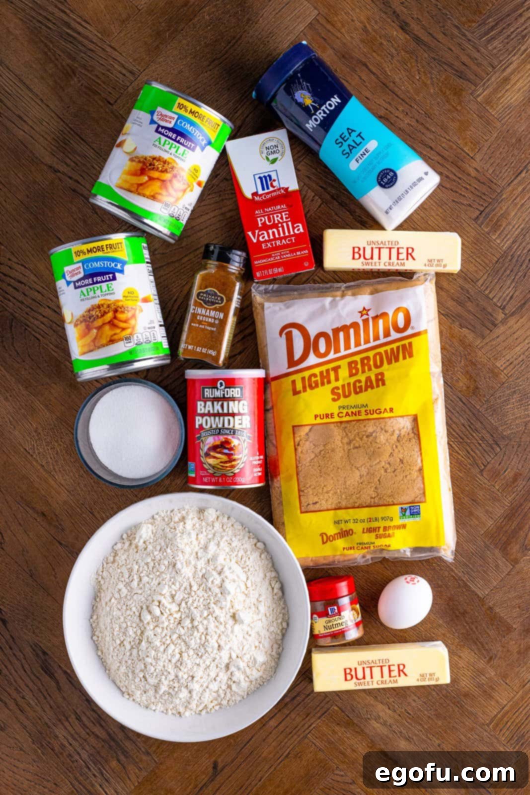 Classic Apple Crumble Bars 4 A flat lay photograph displaying all the ingredients needed for Apple Crumble Bars, including flour, sugars, spices, butter, egg, vanilla extract, and apple pie filling, neatly arranged for preparation.