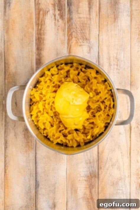 Campus Noodle Comfort 12 Cream of chicken soup being added on top of cooked egg noodles in a pot.