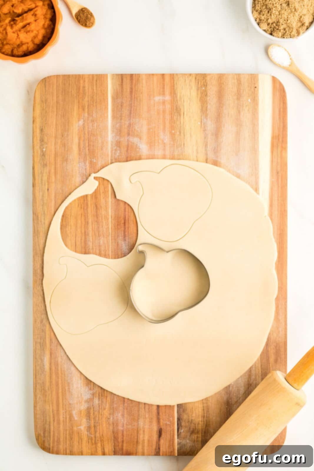 Pumpkin cookie cutter cutting shapes out of pie dough on cutting board.