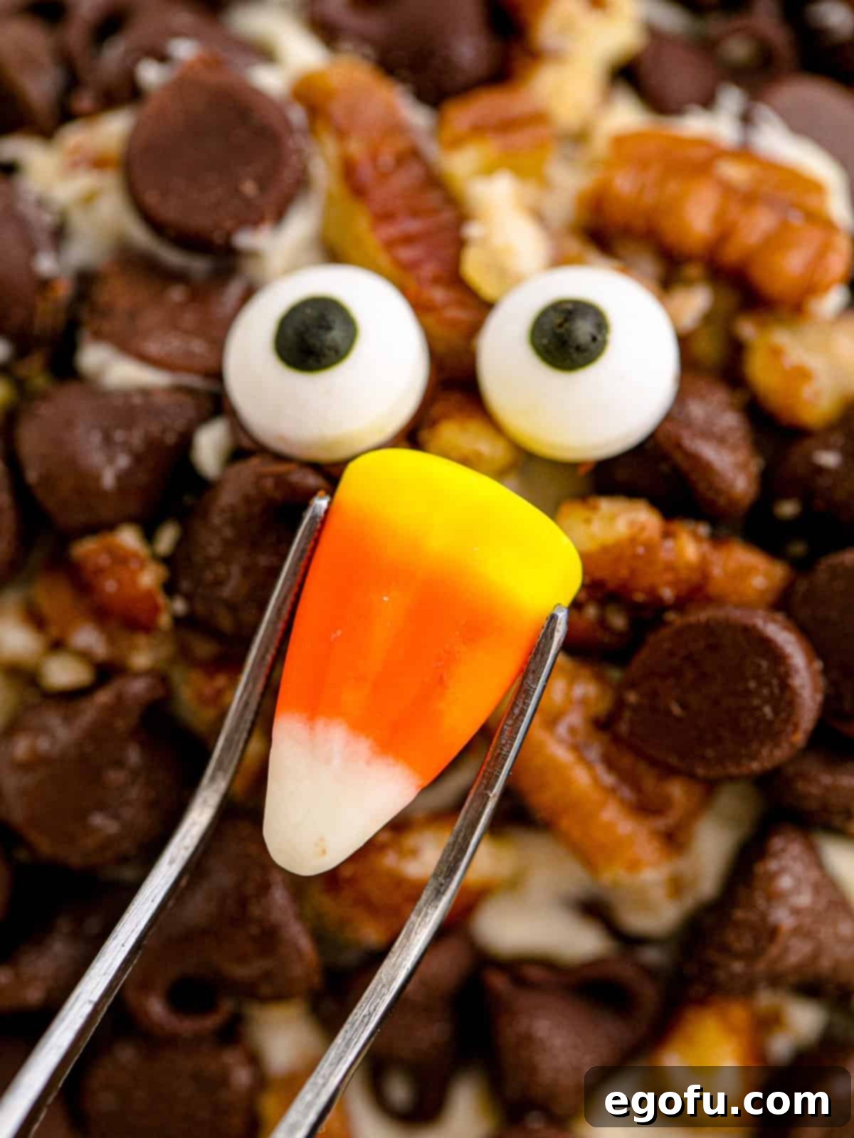 Using tweezers to attach candy eyes and candy corn nose to the front of the cheeseball with melted chocolate.