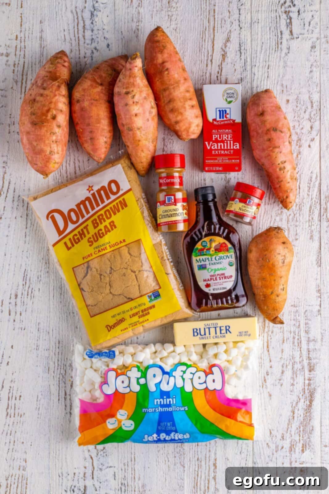 Ingredients needed: sweet potatoes, salted butter, light brown sugar, maple syrup, ground cinnamon, ground nutmeg, vanilla extract, and mini marshmallows, laid out on a surface.