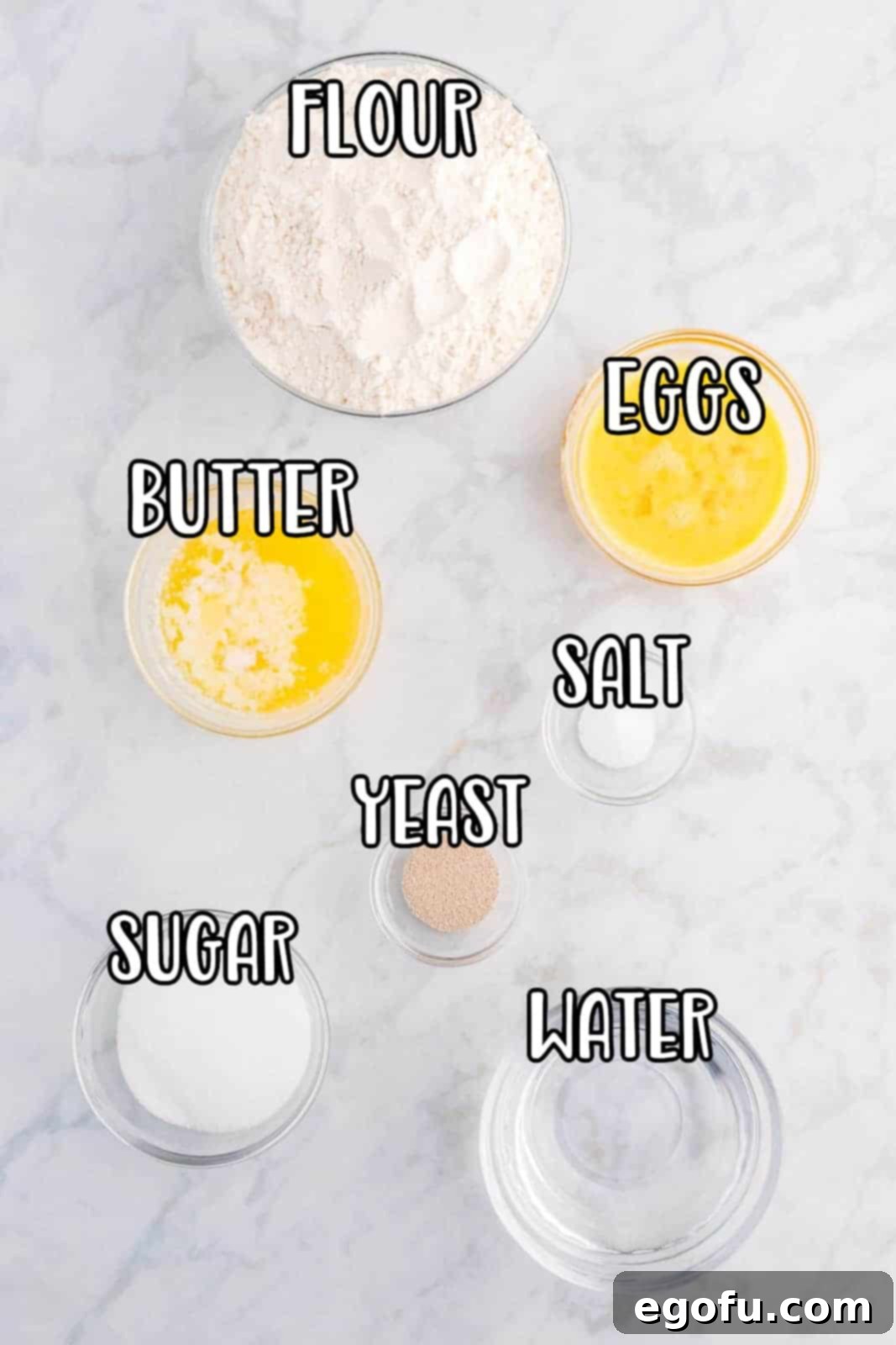 A flat lay photograph showcasing all the essential ingredients for Homemade Crescent Rolls: warm water, active dry yeast, butter, granulated sugar, eggs, salt, and all-purpose flour, arranged neatly on a surface.