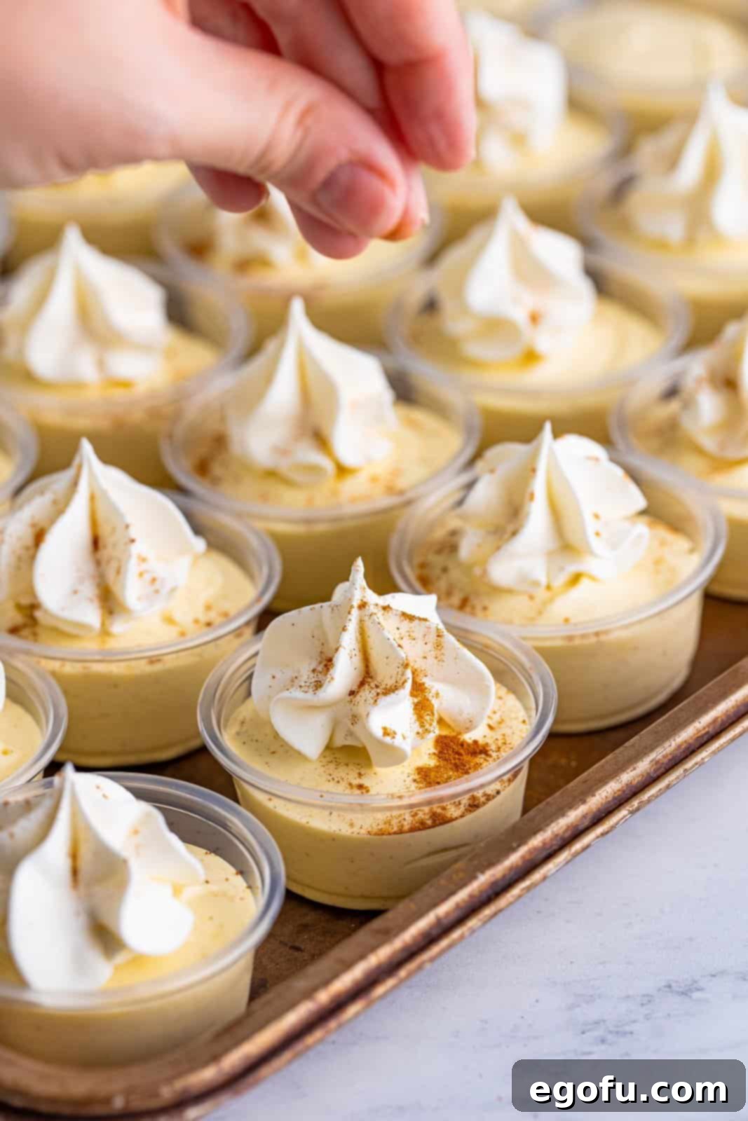 Whipped topping, cinnamon and nutmeg added to tops of pudding shots on sheet tray, highlighting the final presentation.