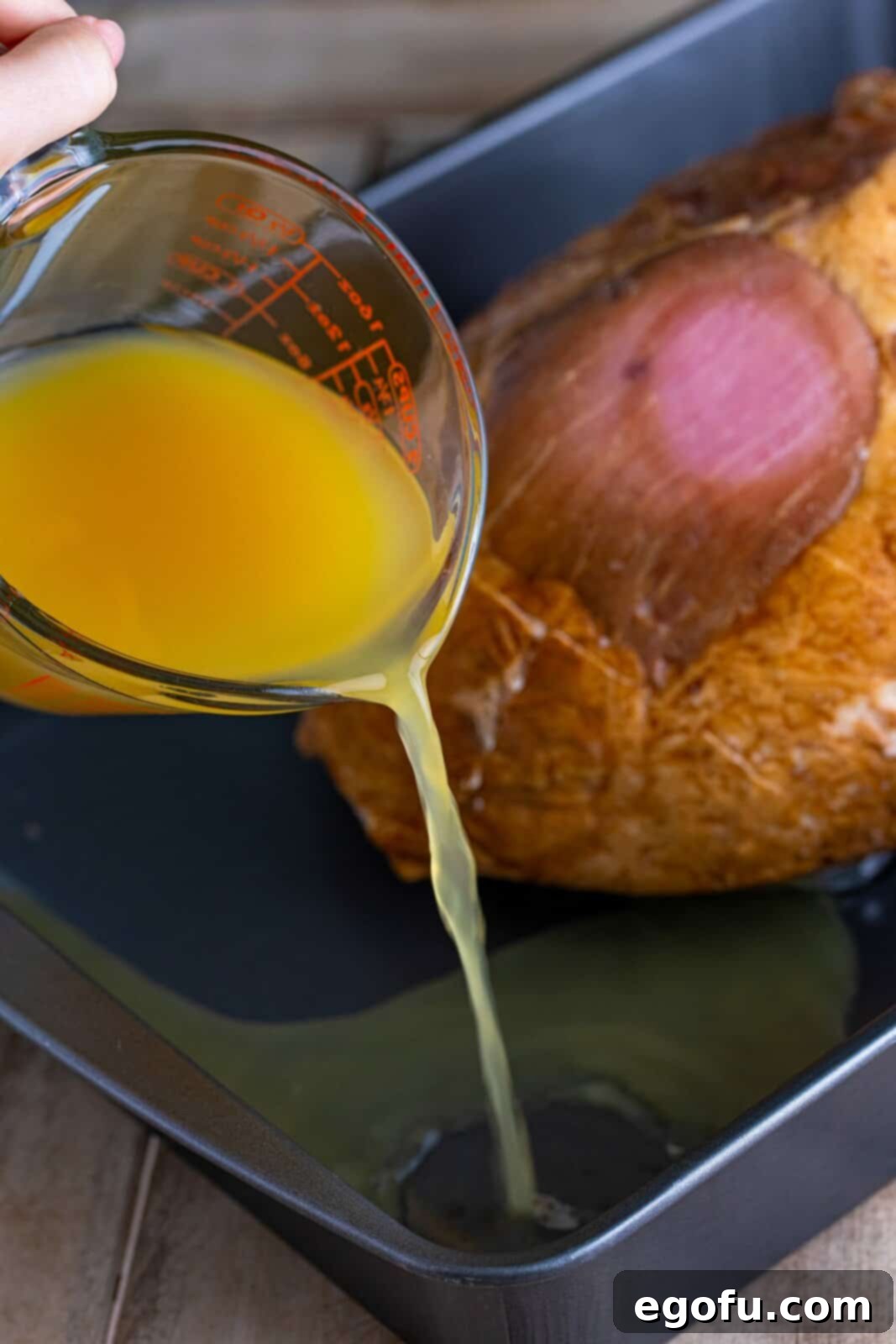 Mahogany Glazed Ham 8 Chicken stock and pineapple juice being poured into the roasting pan around the ham.