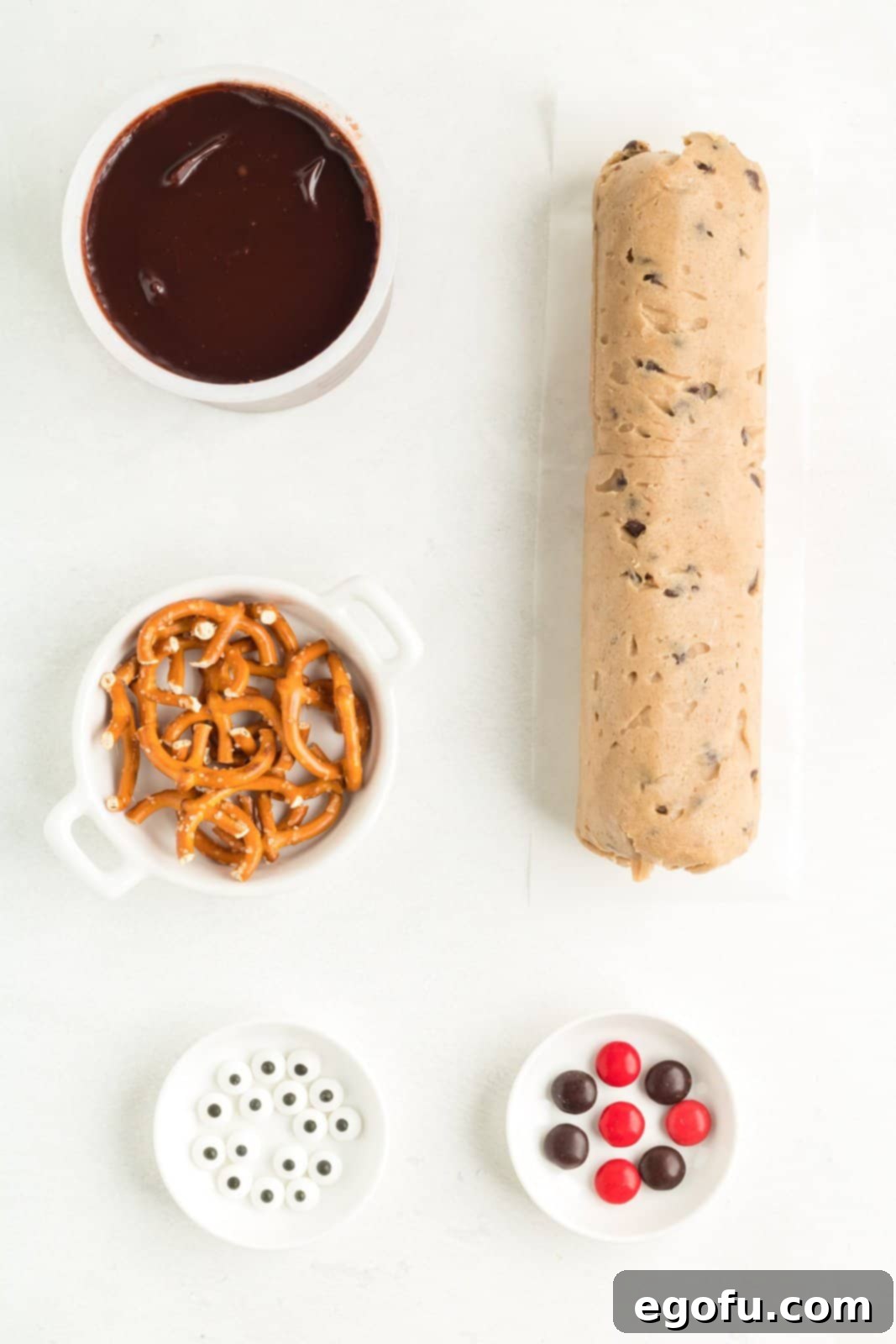 A visual display of all the simple ingredients required to bake Reindeer Cookie Bars: a package of chocolate chip cookie dough, a container of chocolate frosting, a bag of mini pretzel twists, a jar of candy eyes, and a bowl filled with an assortment of red and brown M&Ms.