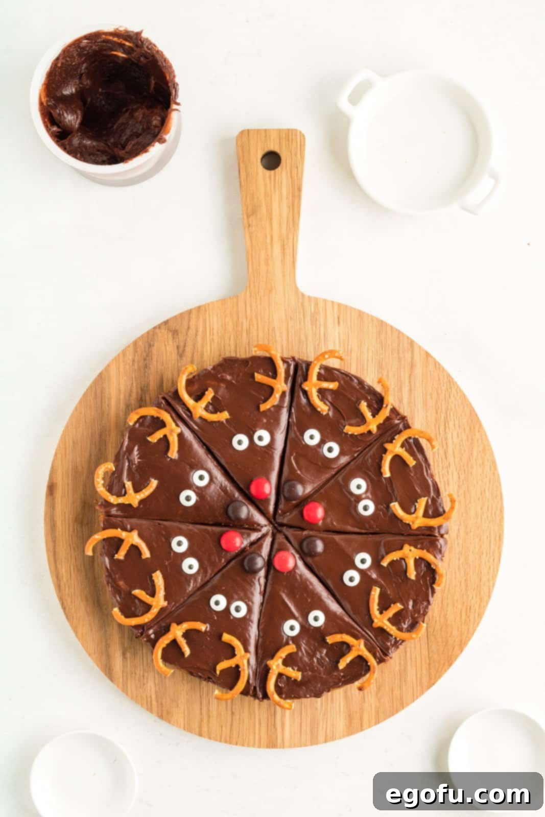 A collection of fully decorated Reindeer Cookie Bars, adorned with pretzel antlers, colorful M&M noses, and whimsical candy eyes, neatly arranged on a round cutting board.
