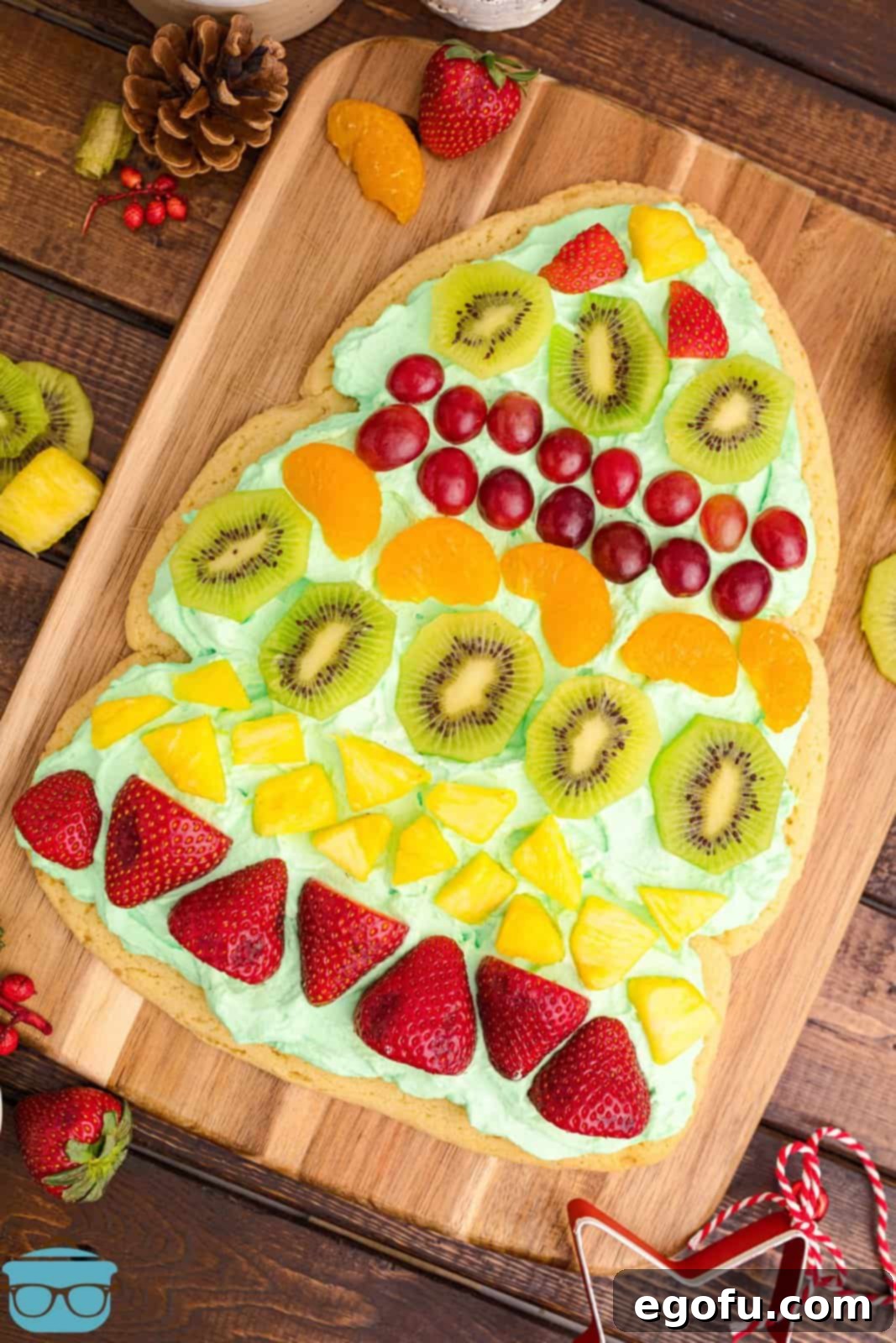 Overhead shot of a vibrant Christmas Tree Fruit Pizza adorned with various fresh fruits on a wooden cutting board, surrounded by additional fruit for decoration.