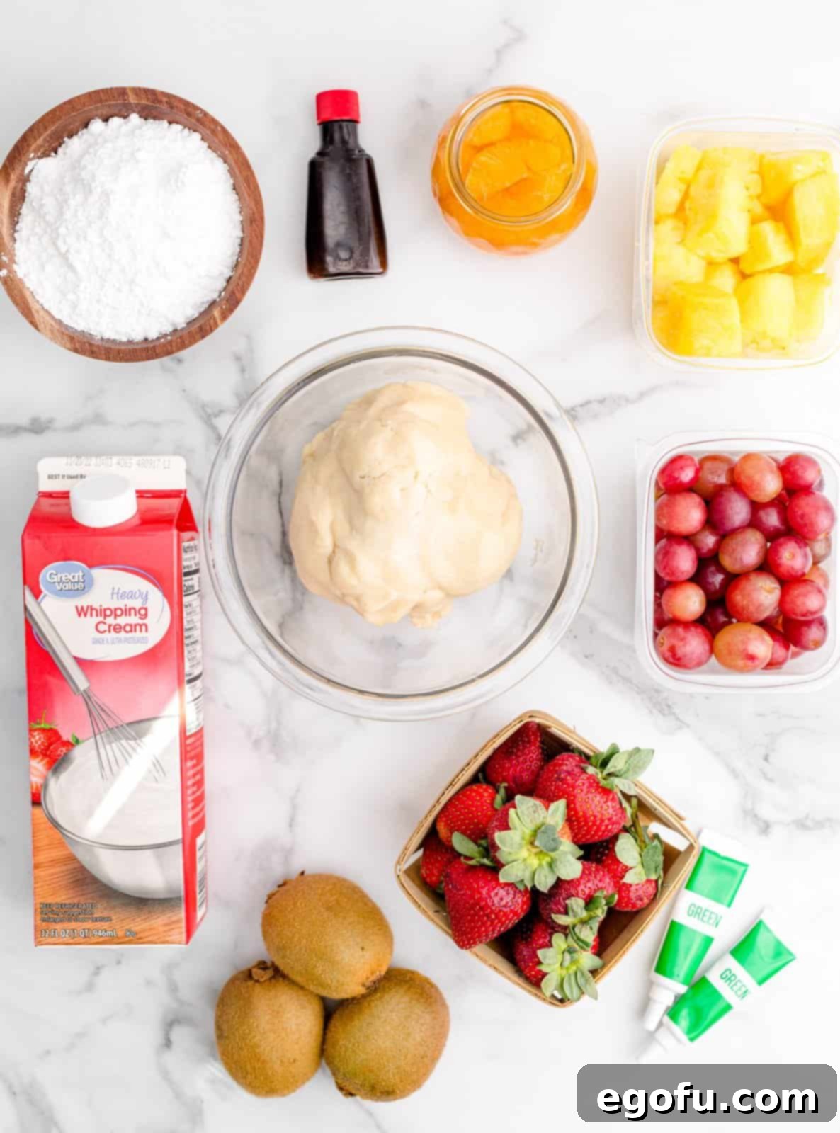 A flat lay photograph of all the ingredients laid out: sugar cookie dough, fresh strawberries, pineapple chunks, green grapes, sliced kiwi, mandarin oranges, heavy cream, powdered sugar, vanilla extract, and green food coloring.