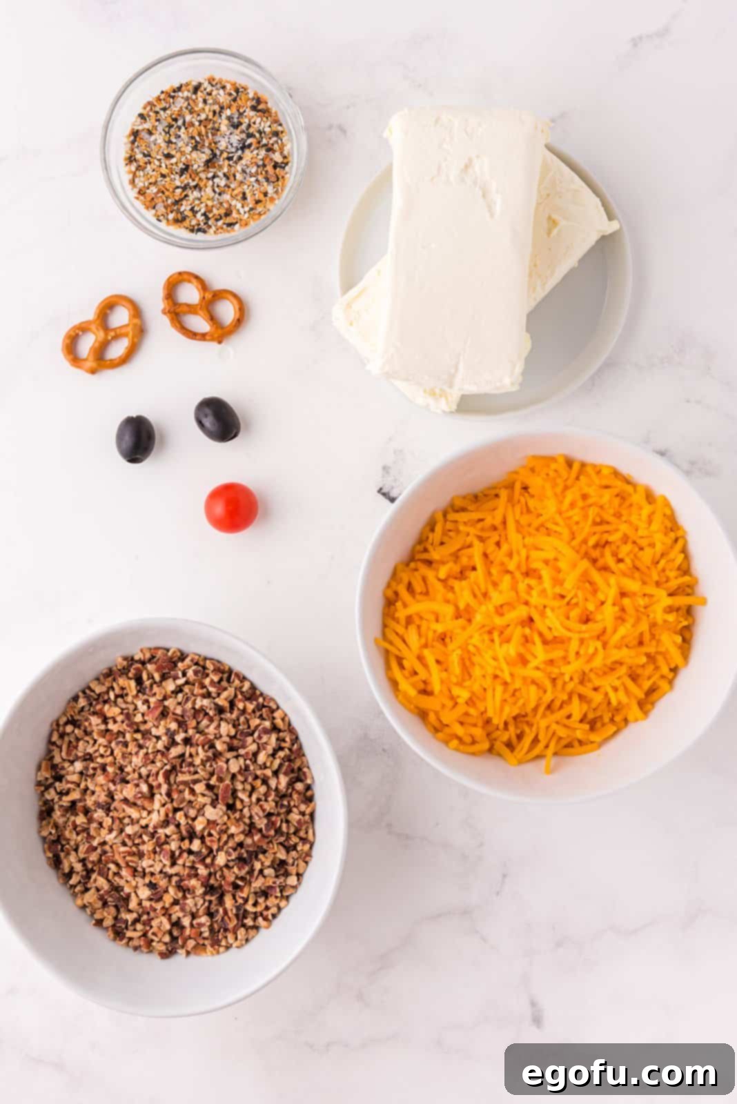 Ingredients for Rudolph Cheeseball: cream cheese, shredded cheddar cheese, everything bagel seasoning, pecans, black olives, cherry tomato, and twisted pretzels.