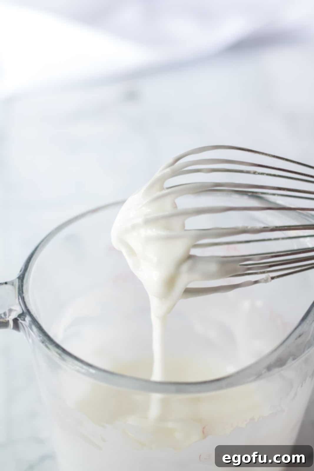 powdered sugar, coffee creamer mixed together in a clear bowl with a whisk holding up some of the icing over the bowl to show thickness. 