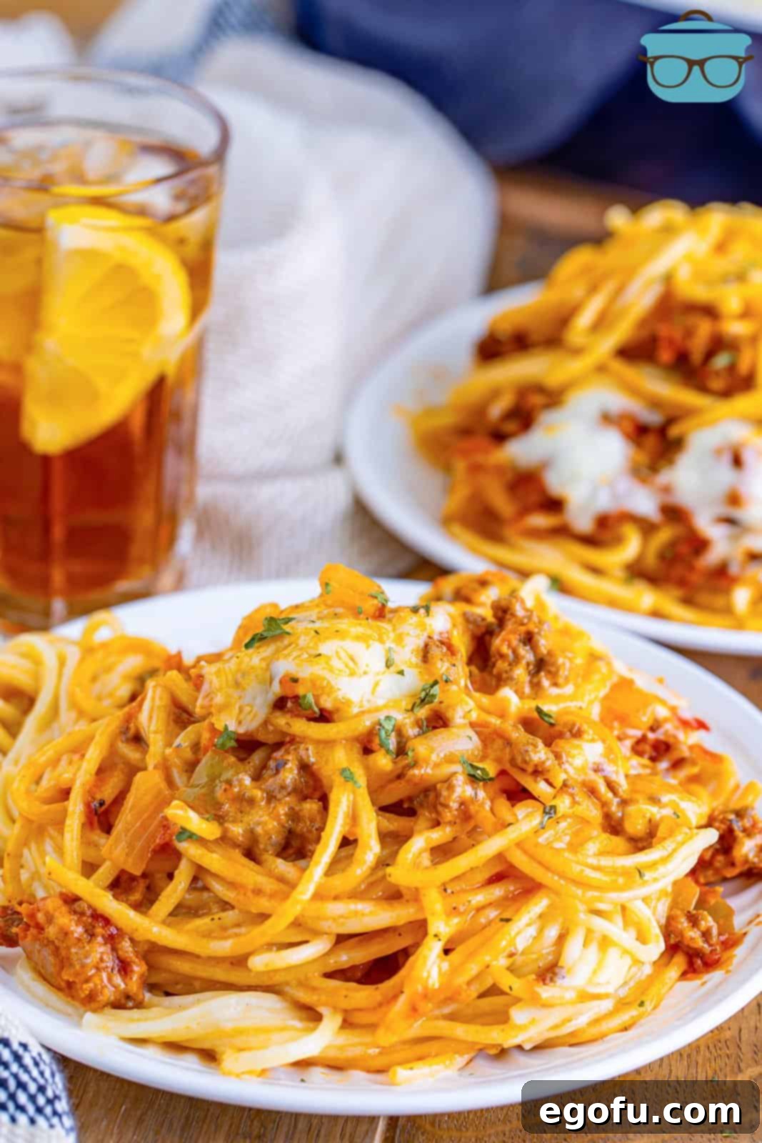 A round white plate with a generous serving of Million Dollar Spaghetti, showcasing its rich layers of pasta, meat sauce, and melted cheese. A glass of iced tea sits in the blurred background, suggesting a delightful meal.
