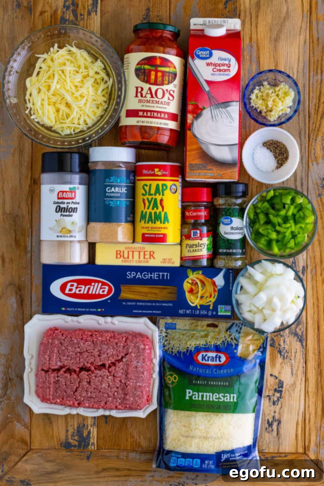 A vibrant flat lay image showcasing all the essential ingredients for Million Dollar Spaghetti: ground beef, onion, bell pepper, various seasonings (cajun, garlic powder, onion powder, Italian), marinara sauce, unsalted butter, fresh garlic, heavy cream, uncooked spaghetti noodles, and a selection of shredded cheeses.