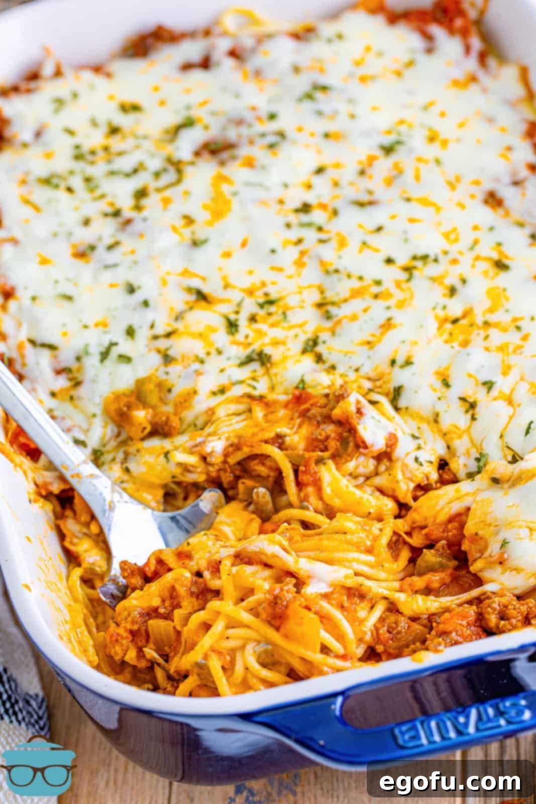A close-up shot of the finished Million Dollar Spaghetti casserole, with a large silver spoon inserted, ready to serve a portion. The melted, bubbly cheese topping and layers of sauce are clearly visible.