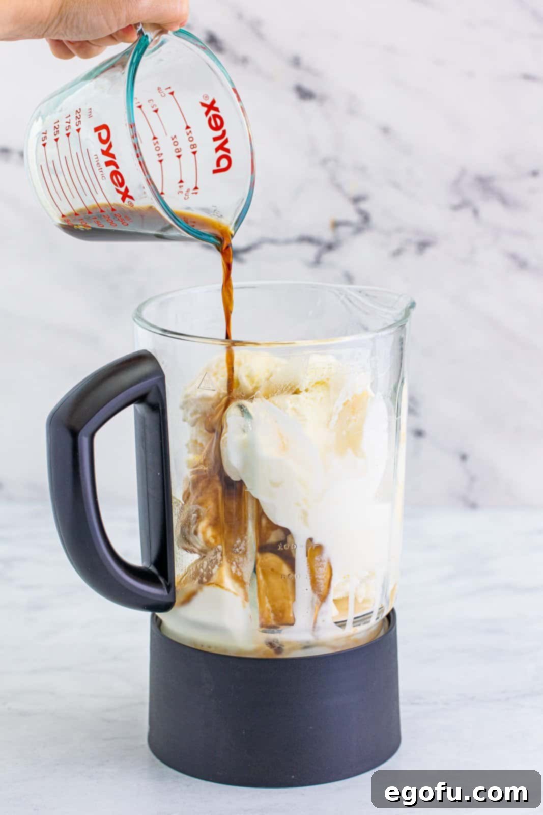 Rudolph on the Rocks 5 A hand pouring coffee liqueur into a blender filled with vanilla ice cream, as part of the Drunken Rudolph milkshake preparation.