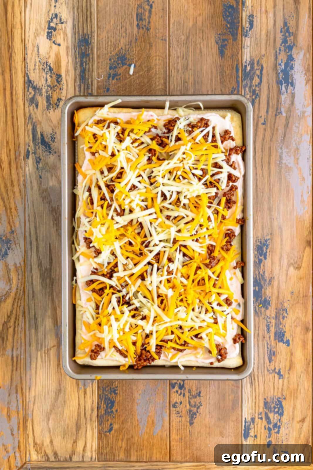 Shredded cheddar and Monterey Jack cheeses evenly sprinkled on top of the taco pizza layers, ready for baking.
