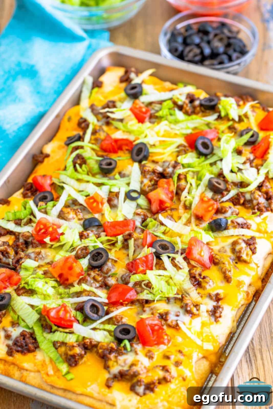 Taco pizza shown in a baking sheet being topped with fresh shredded lettuce, diced tomatoes, and sliced olives.