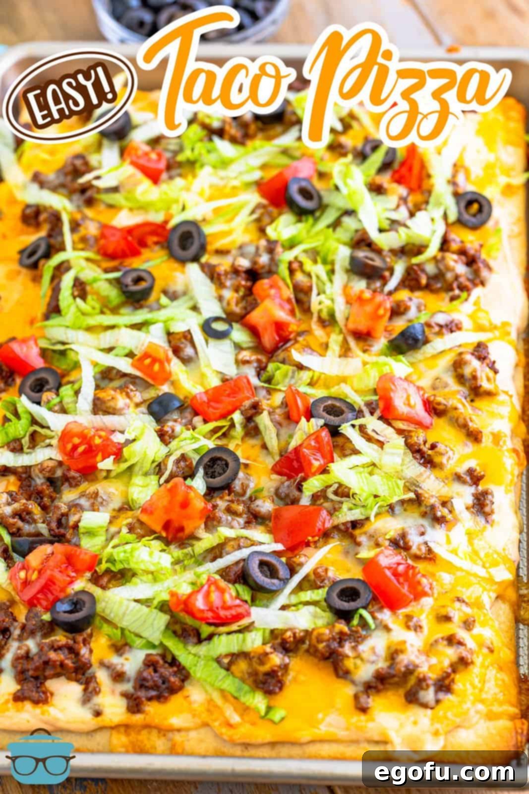 A fully made beef taco pizza shown in a baking pan, generously topped with cheese and ready for fresh additions.