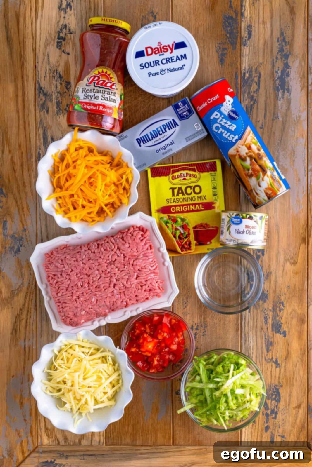 Individual ingredients for Taco Pizza: pizza dough, ground beef, taco seasoning, water, cream cheese, salsa, sour cream, and shredded cheese, laid out on a surface.