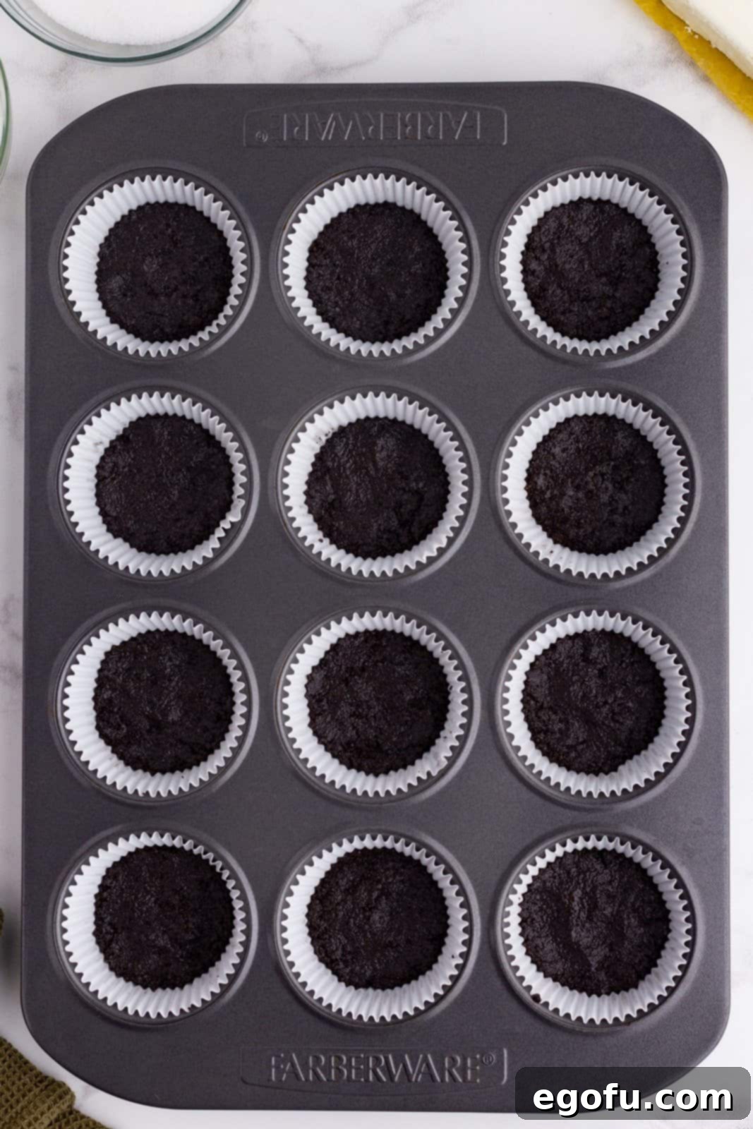 Oreo cookie crumb mixture shown pressed into the bottom of cupcake liners in a pan.