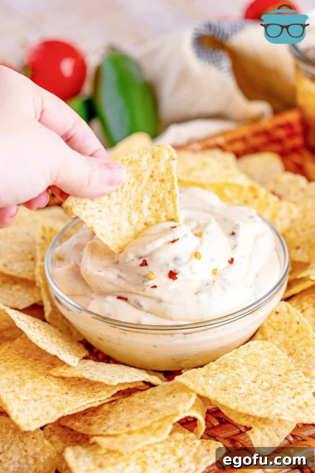 A hand holding a golden tortilla chip, dipping it into a small, clear glass bowl filled with creamy Virginia White Sauce.