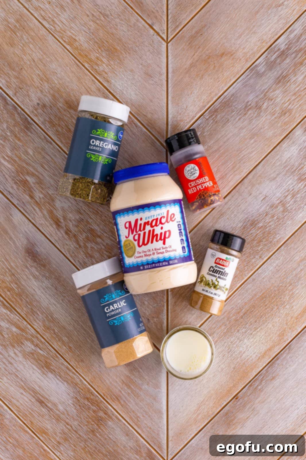Individual ingredients laid out: Miracle Whip jar, a small bowl of dried oregano, garlic powder, crushed red pepper flakes, ground cumin, and a glass of milk.