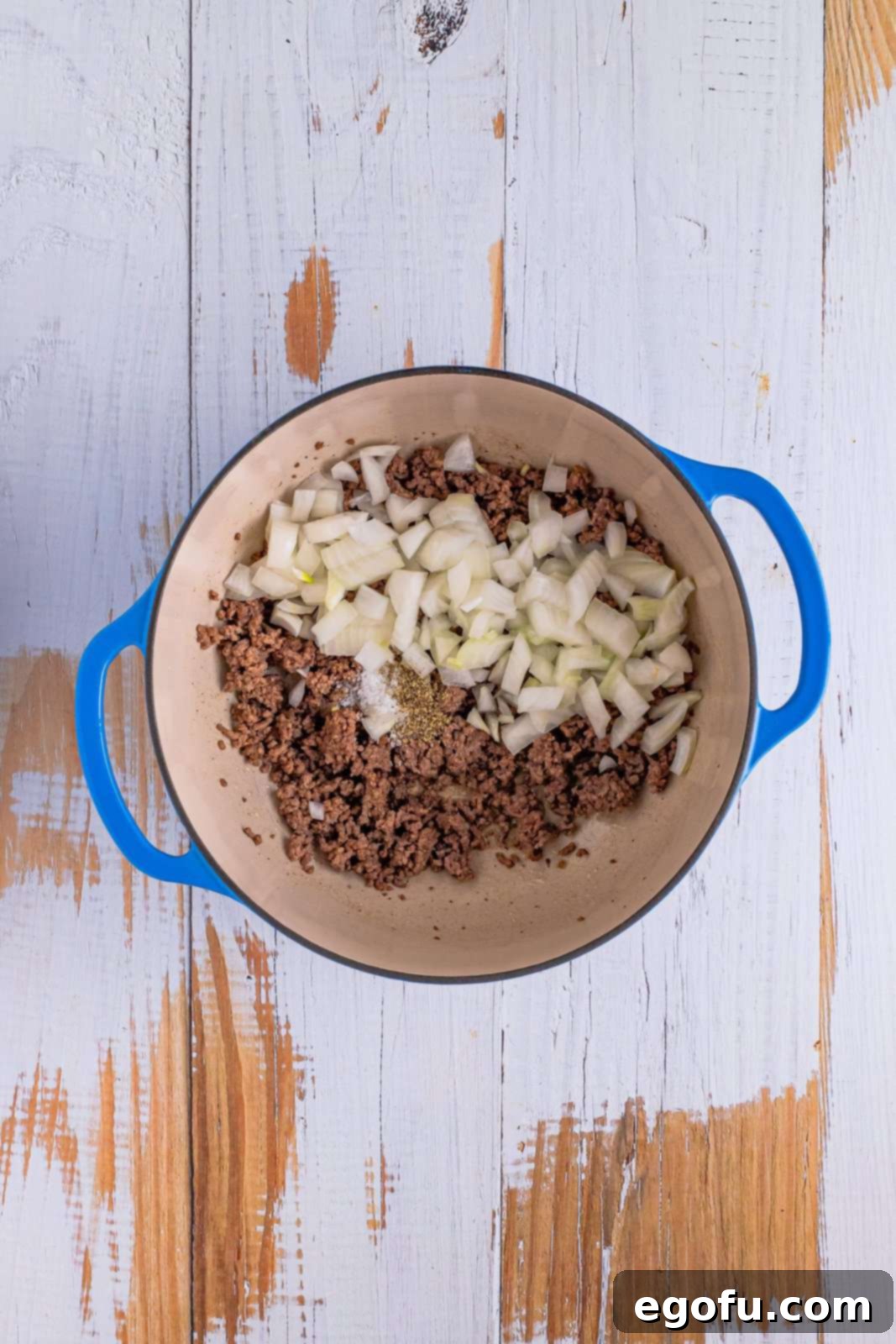A dutch oven with ground beef and raw onion on top with salt and pepper.