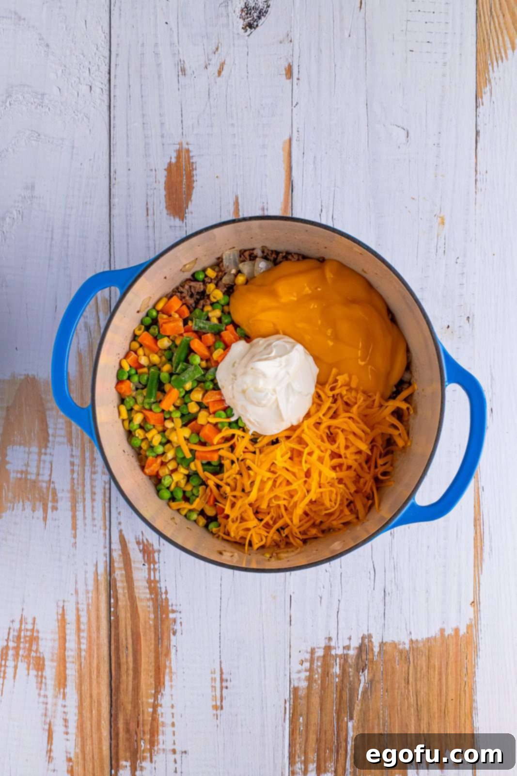 Looking down on a dutch oven with ground beef, onion, herbs spices and cheddar, mixed veggies, and cream of cheddar soup on top.