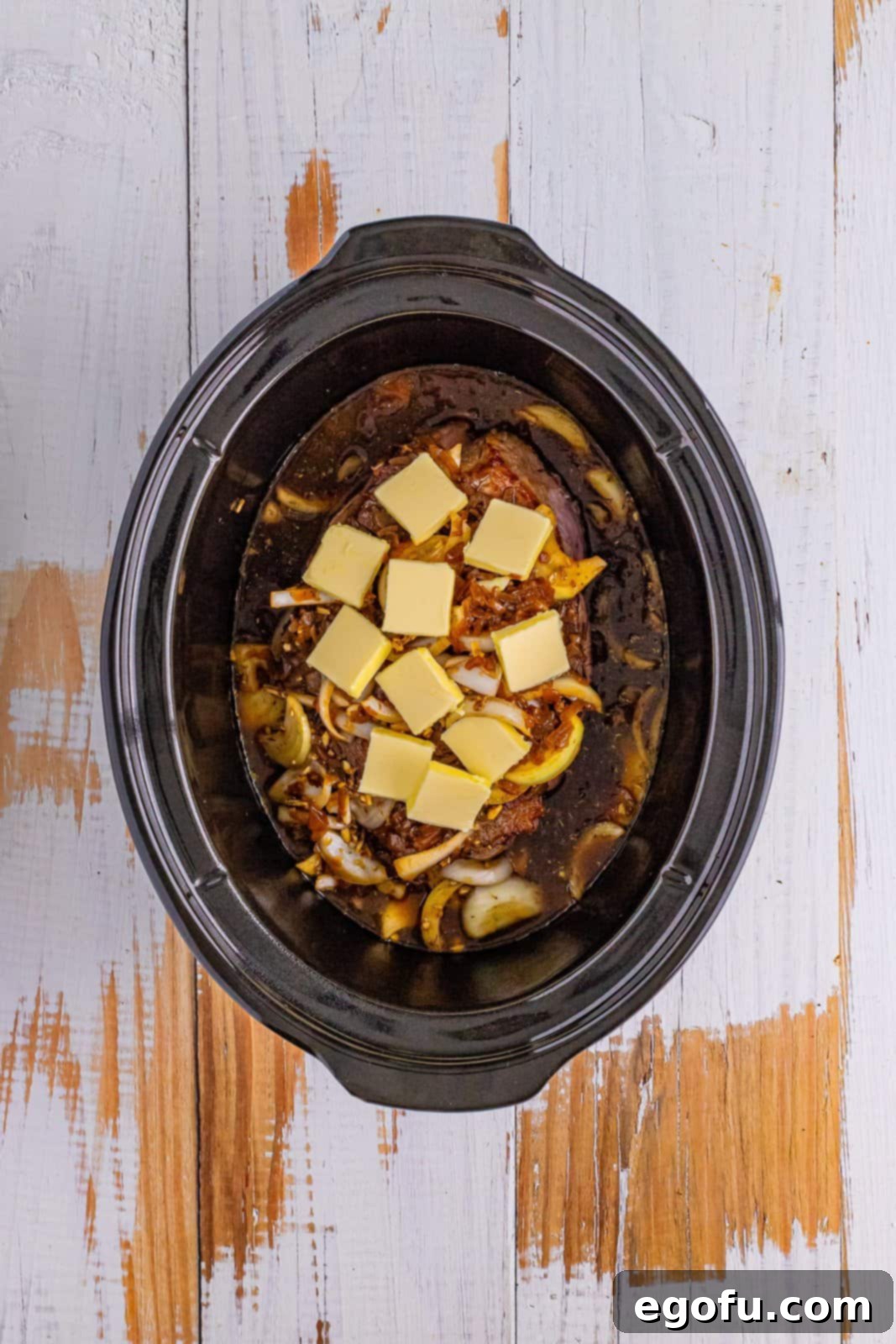 Looking down on a Slow Cooker filled with pot roast, condensed soup, onions, spices, and melting sliced butter, ready for slow cooking.