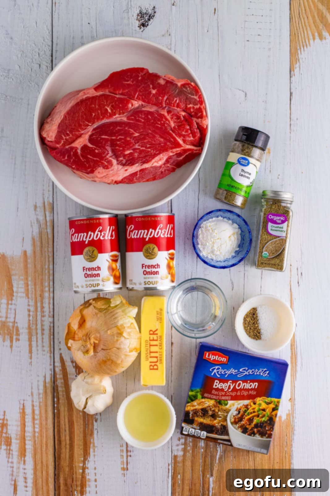A collection of ingredients laid out on a table: an onion, garlic, beefy onion soup mix packet, dried thyme, dried rosemary, cans of condensed french onion soup, a chuck roast, kosher salt, black pepper, vegetable oil, unsalted butter, water, and cornstarch.