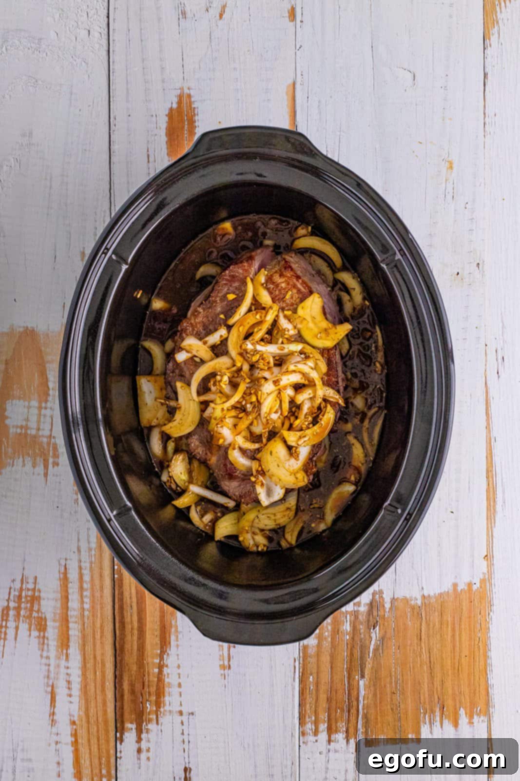 Pot roast nestled in the oval slow cooker, topped with remaining onions, condensed soup, and slices of butter.