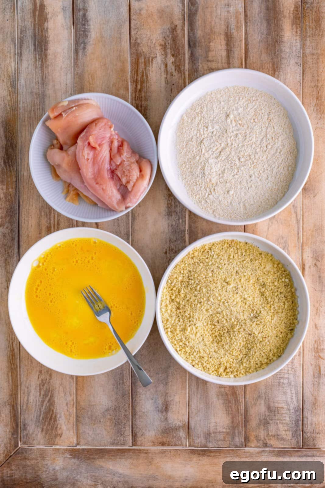 Four bowls, one with chicken breasts, one with a breadcrumb mixture, one with egg mixture and one with flour and spices. 