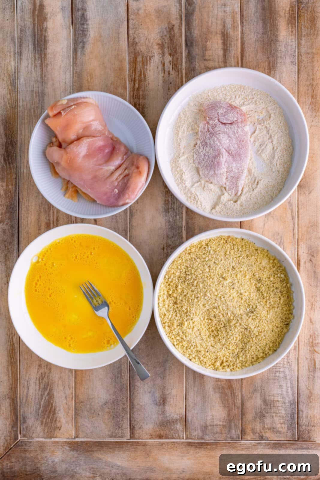 Four bowls- one with chicken, one with a piece of chicken in the flour mixture, another bowl with panko mixture and a final bowl with egg mixture. 