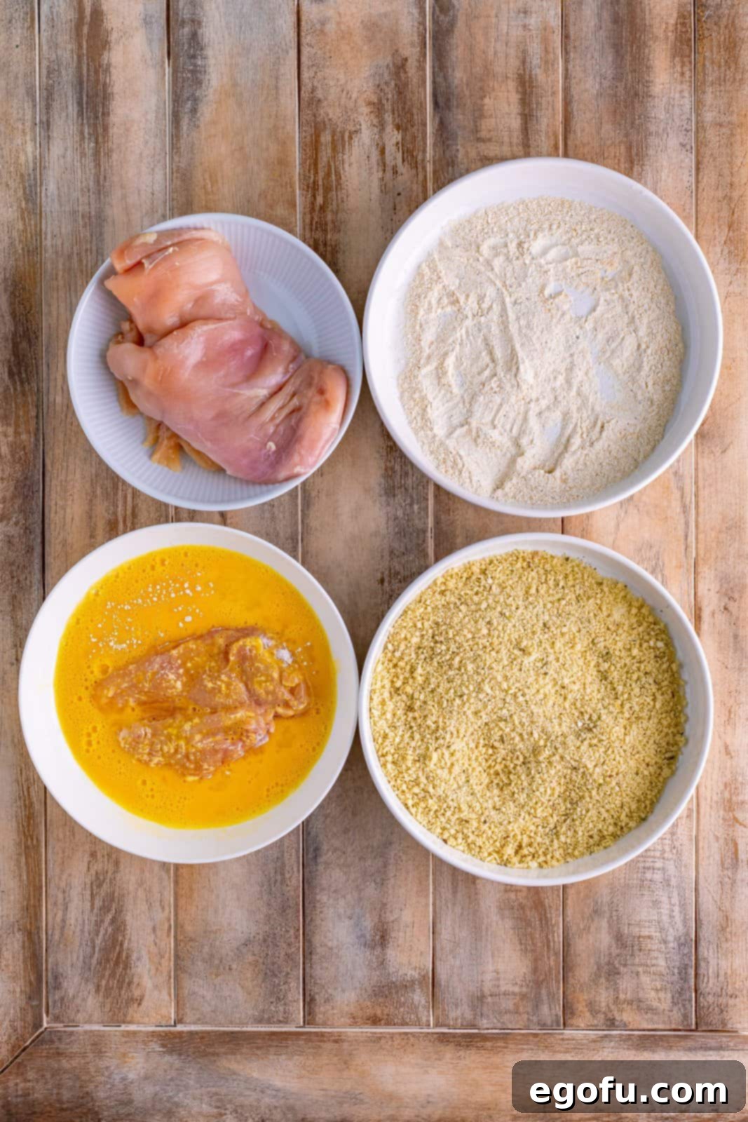 4 Bowls of different breading ingredients with chicken in egg mixture.