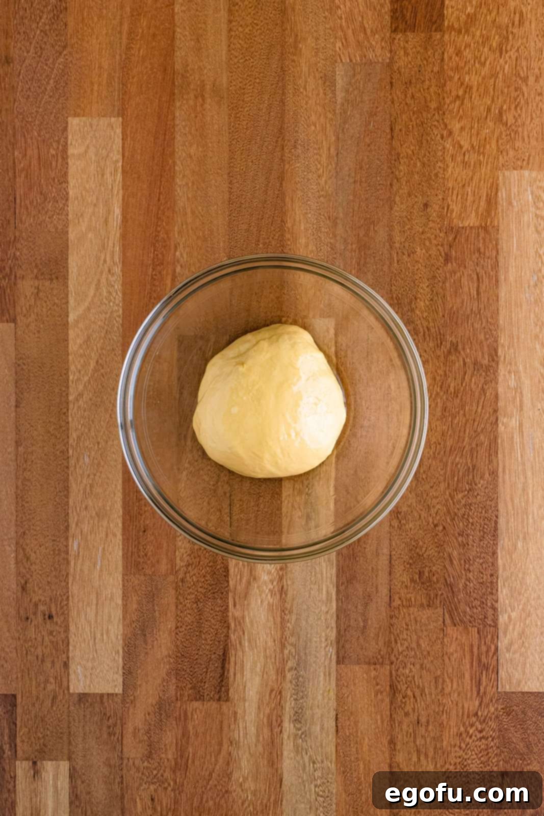 Butter Bathed Cloud Rolls 10 Glass bowl containing dough that has successfully risen and doubled in size, covered with plastic wrap.