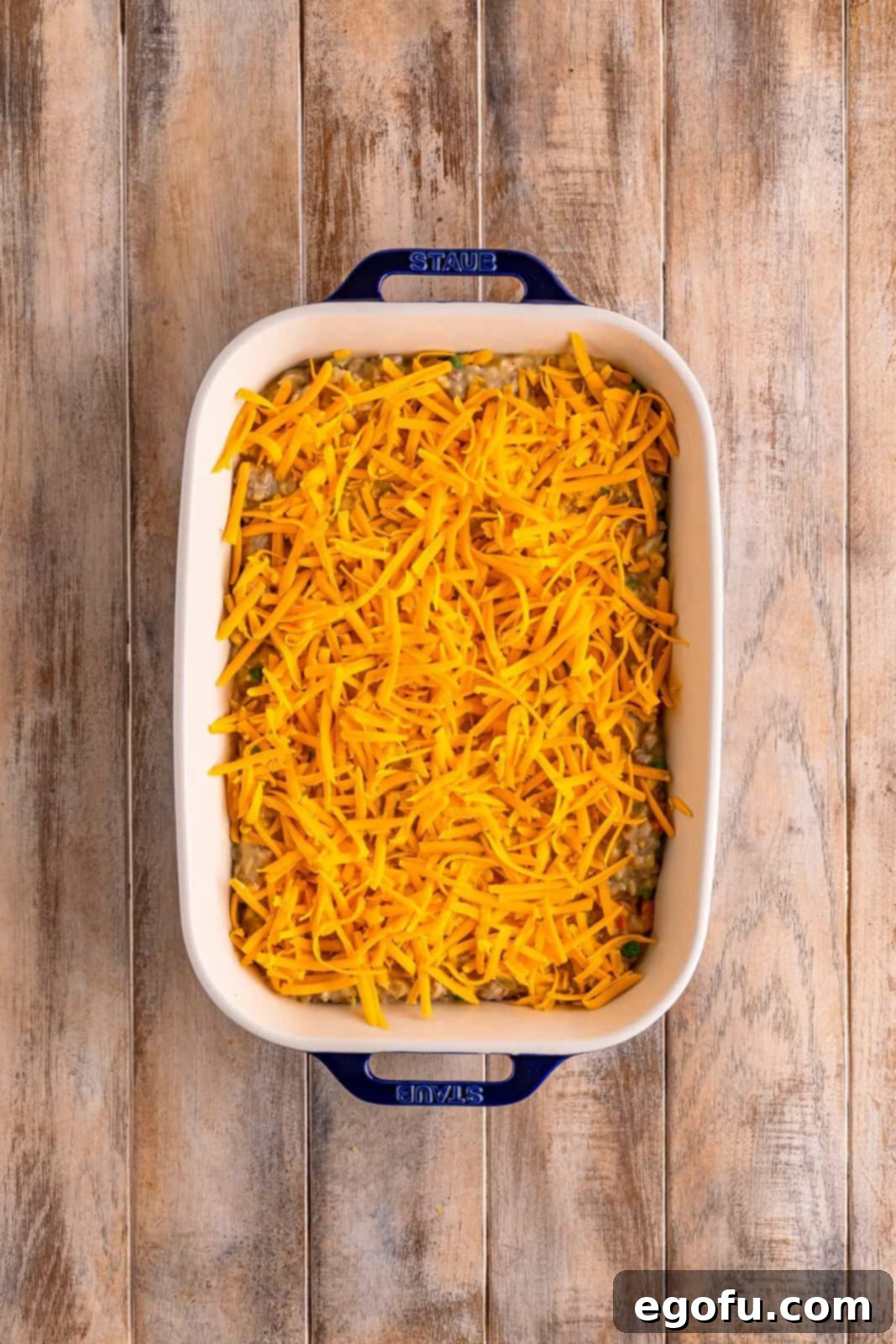 Golden Cheesy Beef and Rice Bake 8 A casserole dish with an uncooked ground beef and rice mixture covered with a generous layer of shredded cheddar cheese, ready to be baked.