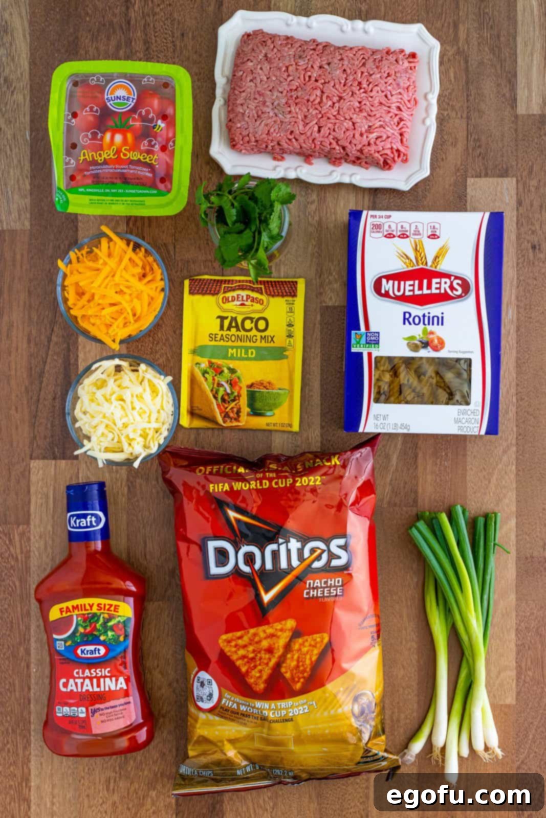 A flat lay of all the ingredients for Taco Pasta Salad: Rotini pasta, ground beef, taco seasoning, cherry tomatoes, scallions, shredded sharp cheddar cheese, shredded Monterey Jack cheese, Catalina dressing, crushed nacho cheese Doritos, and fresh cilantro.