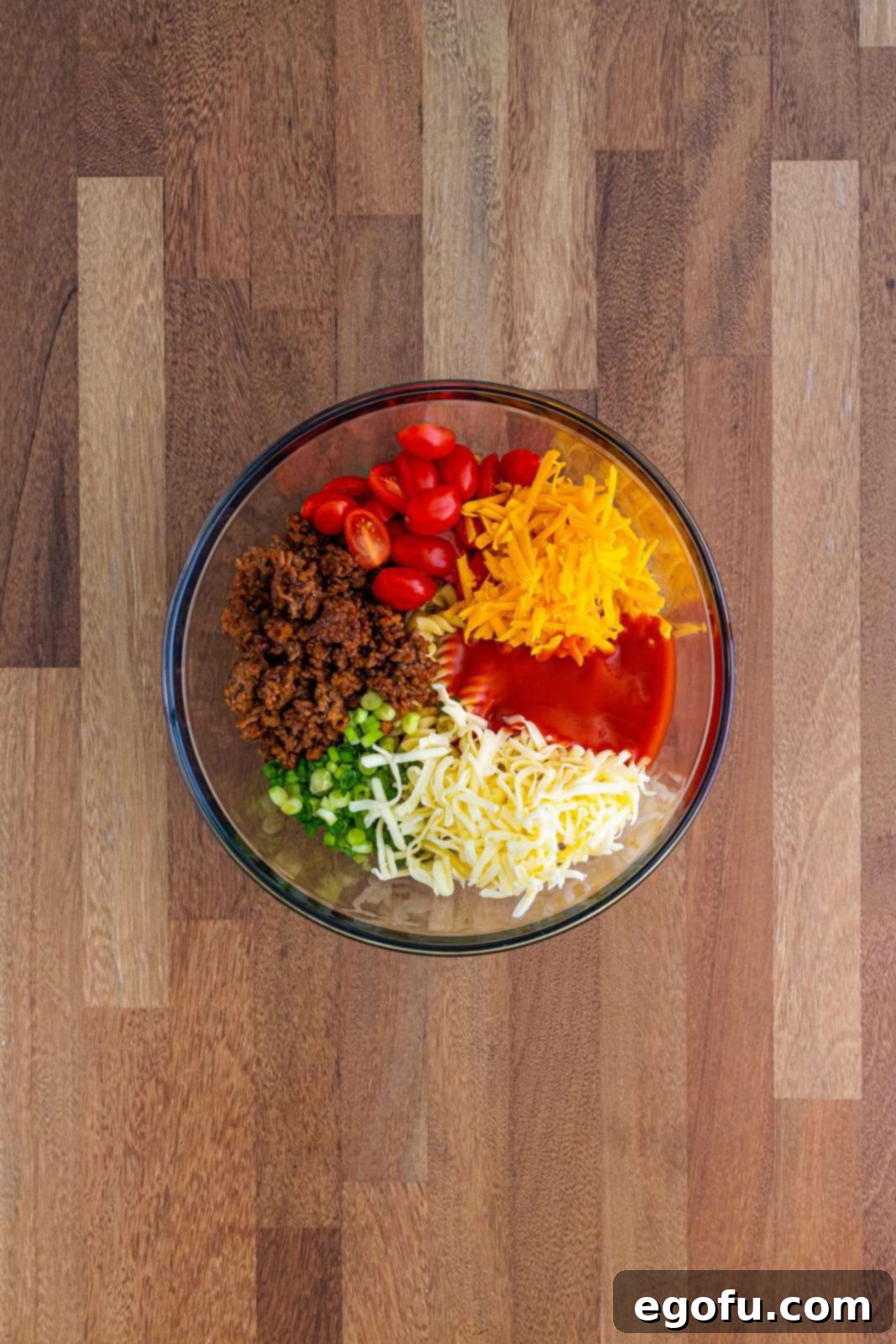 A large bowl filled with pasta, ground beef mixture, cherry tomatoes, scallions, cheddar cheese, Monterey Jack cheese, and Catalina dressing, ready for mixing.