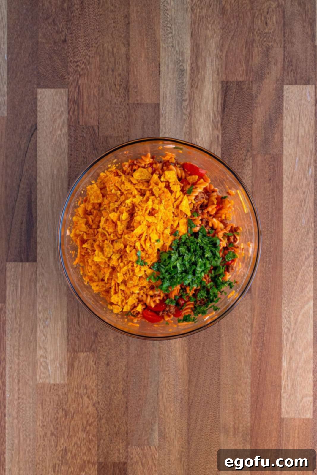 A glass bowl with Taco Pasta salad and crushed Doritos and cilantro being mixed in.