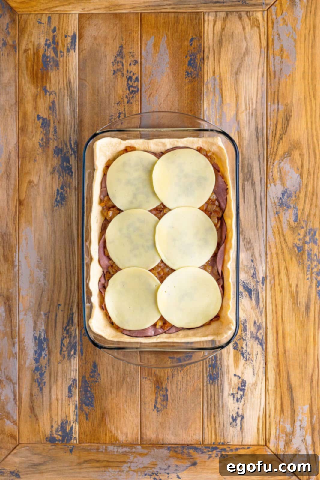 A baking dish perfectly layered with crescent dough, provolone cheese, roast beef, caramelized onion, and topped with the final layer of provolone cheese.
