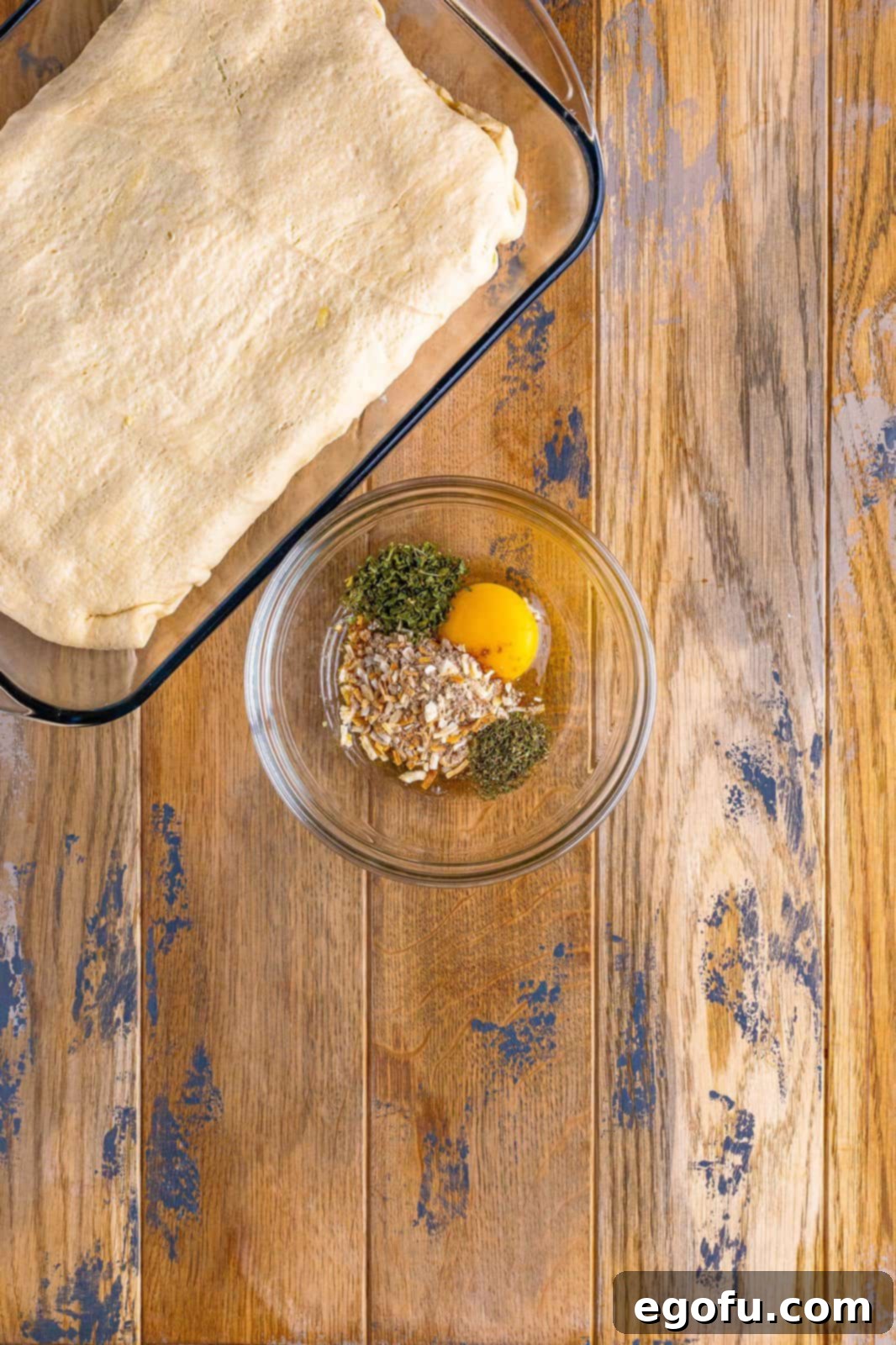 A small glass mixing bowl containing a whisked egg, dried onion soup mix, parsley flakes, and thyme, ready to be brushed onto the dough.