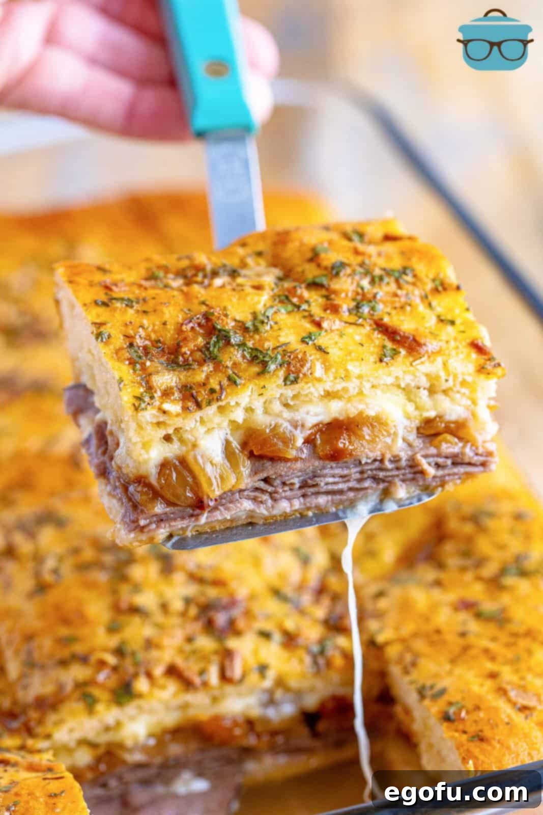 A serving utensil holding a perfectly cut French Dip Square above a baking dish filled with more servings, ready to be enjoyed.
