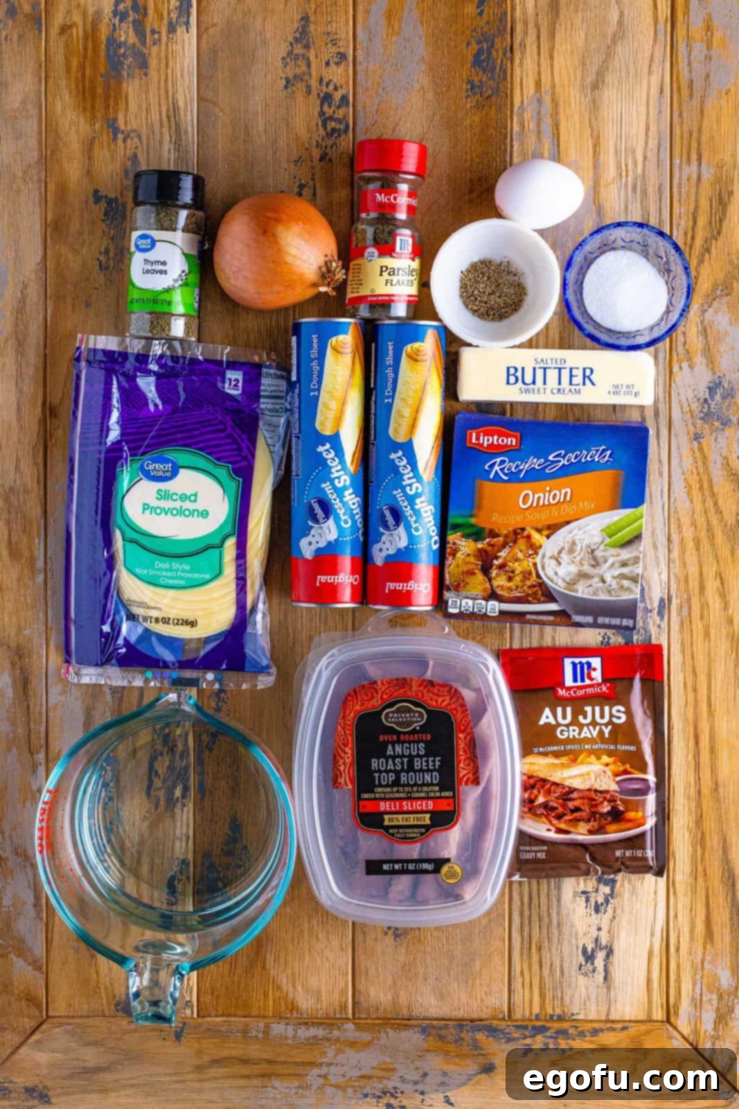 Arrangement of crescent dough sheets, delicious sliced roast beef, au jus gravy packet, onion soup mix, salted butter, provolone cheese, onion, egg, dried thyme and dried parsley flakes, illustrating the ingredients for French Dip Squares.